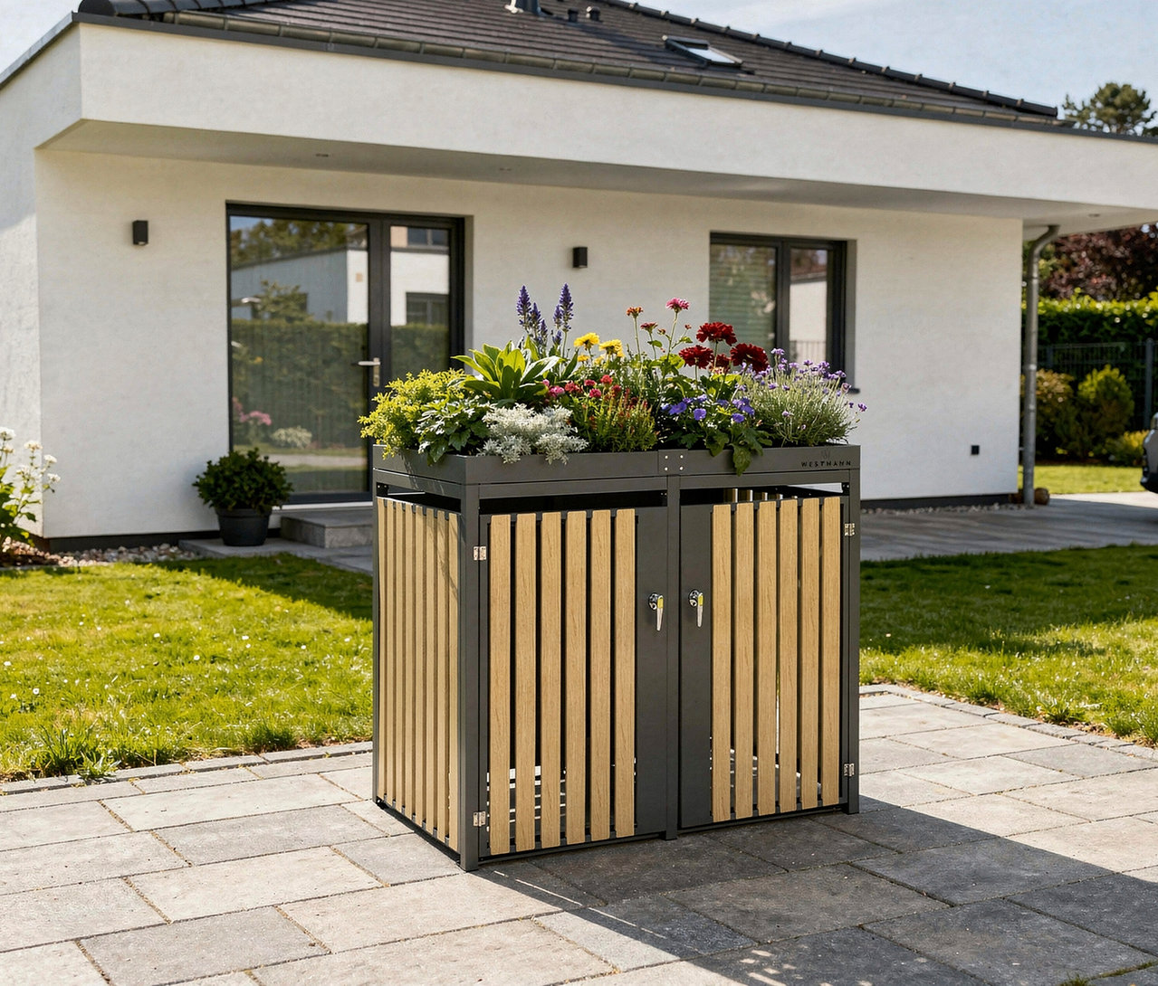 Eine Mülltonnenbox mit Blumenkasten für 2 Tonnen steht vor einem Haus auf einem gepflasterten Weg.
