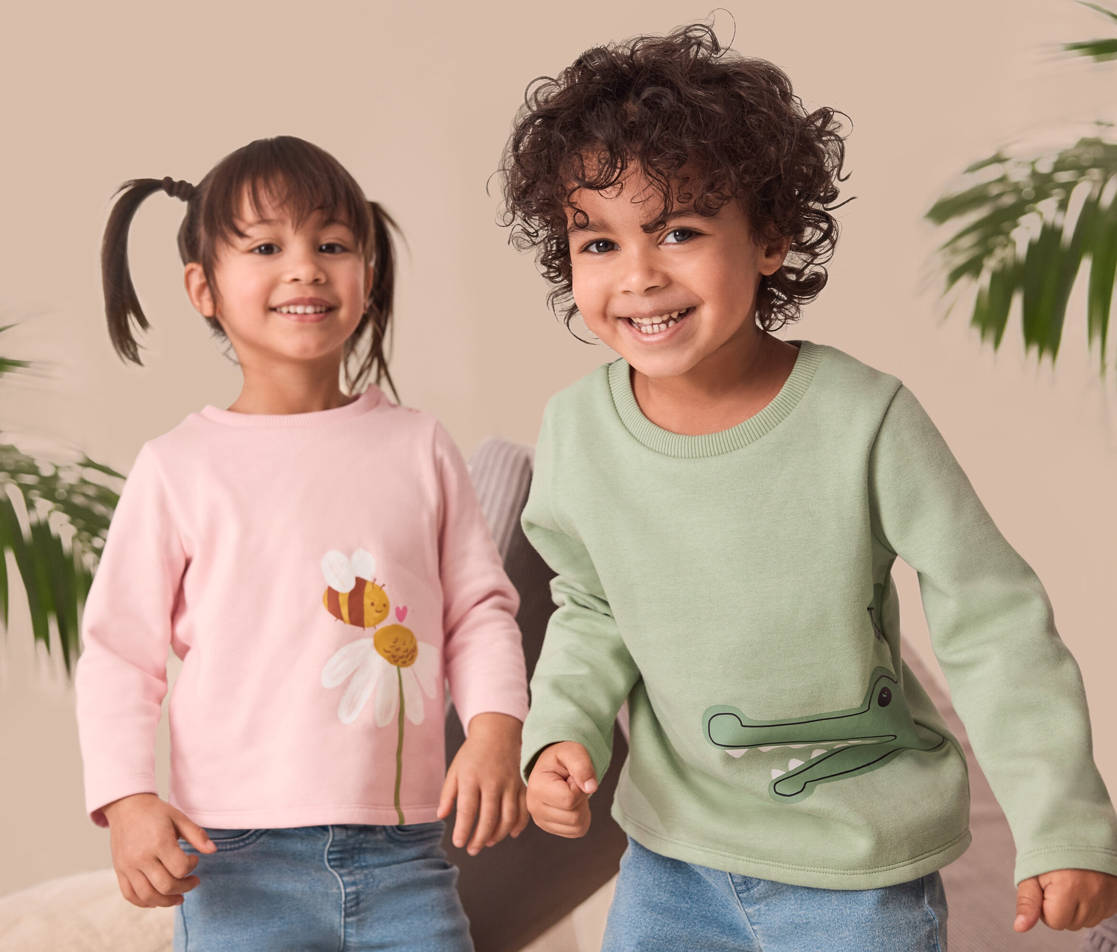 Zwei lächelnde Kinder stehen nebeneinander. Das Mädchen links trägt ein rosa Kinder-Sweatshirt mit einem Blumen- und Bienen-Print. Der Junge rechts trägt ein grünes Kinder-Sweatshirt mit Krokodil-Print.