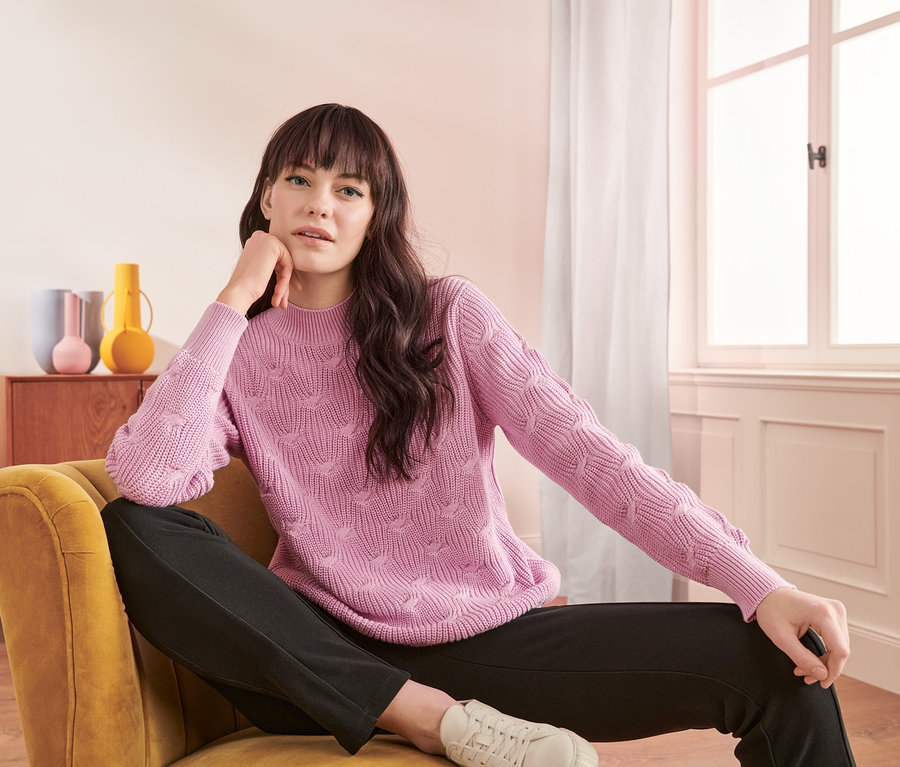 Frau sitzt im Sessel und trägt einen Pullover mit Zopfmuster.
