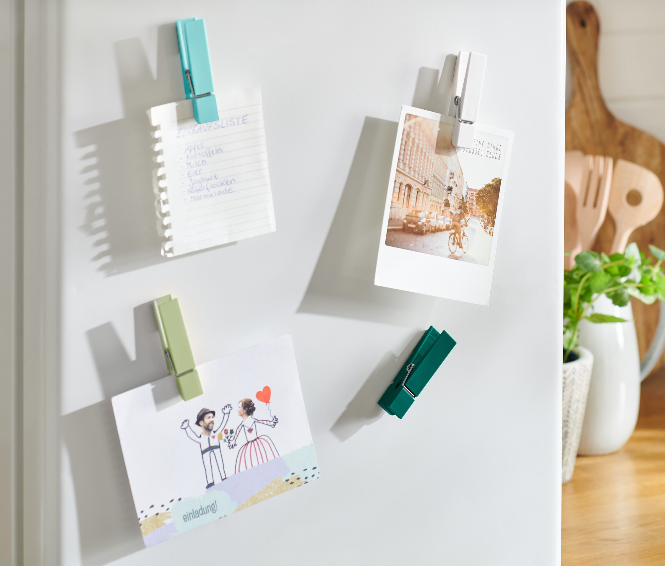 Vier Kühlschrankmagnete halten eine Einkaufsliste, eine Postkarte und eine Einladung.