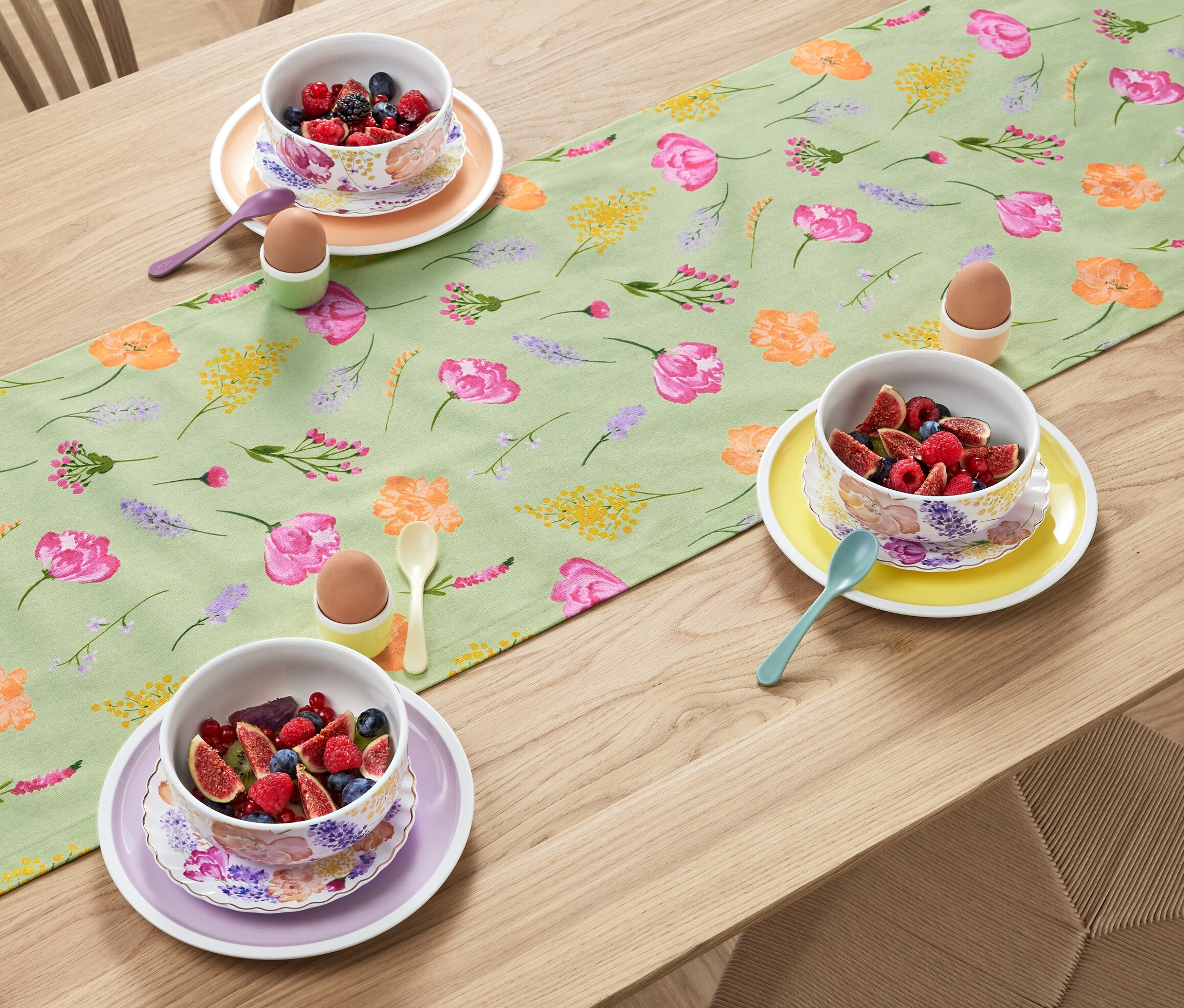 Auf einem Holztisch liegt ein salbeifarbener Tischläufer mit Blumendruck, auf dem Schüsseln mit Obst, Eier in Haltern und Löffel stehen.