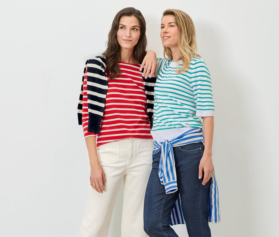 Zwei Frauen posieren vor einem weißen Hintergrund. Eine trägt einen gestreiften Feinstrickpullover in Aqua und Jeans, die andere einen rot-weiß gestreiften Pullover und eine weiße Hose.