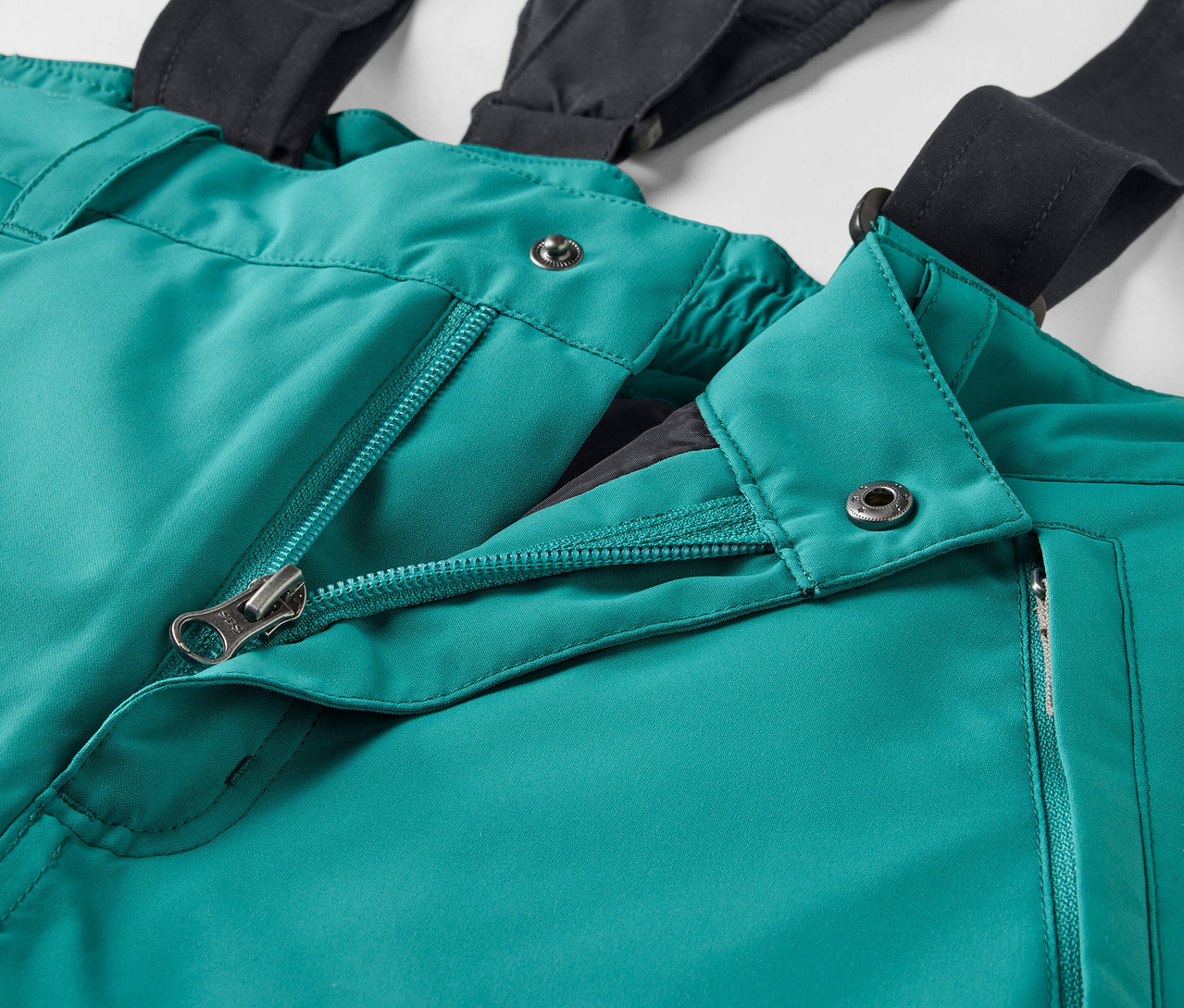 Detail einer grünen Skihose mit schwarzen Hosenträgern und offenem Reißverschluss.