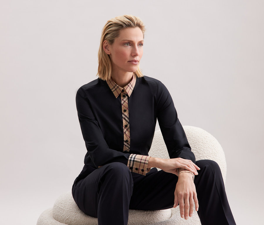 Blonde Frau sitzt auf einem weißen Stuhl und trägt schwarze Kleidung mit Karomuster.