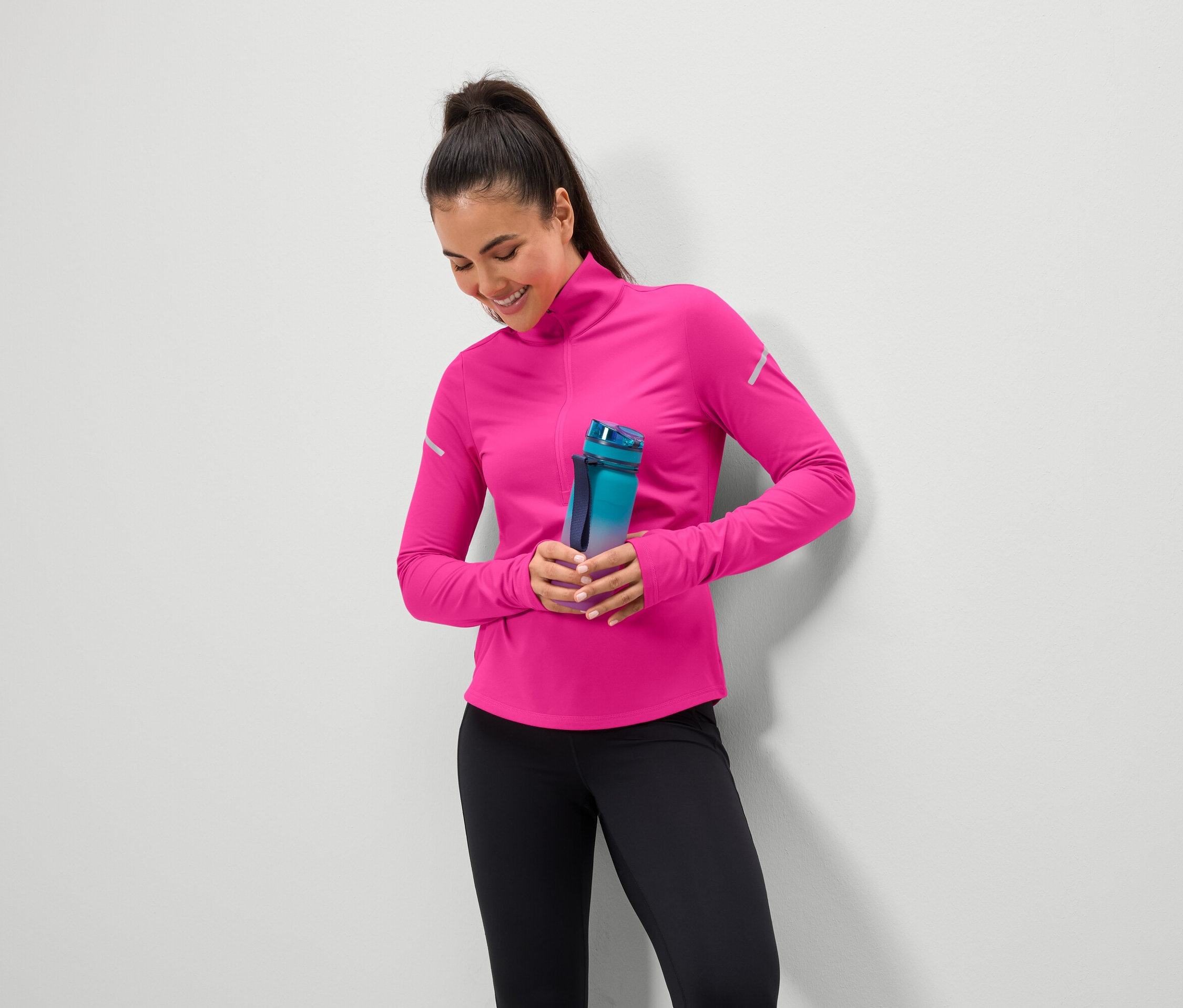 Lächelnde Frau hält eine blaugrüne Wasserflasche, trägt ein pinkes Oberteil und schwarze Leggings.