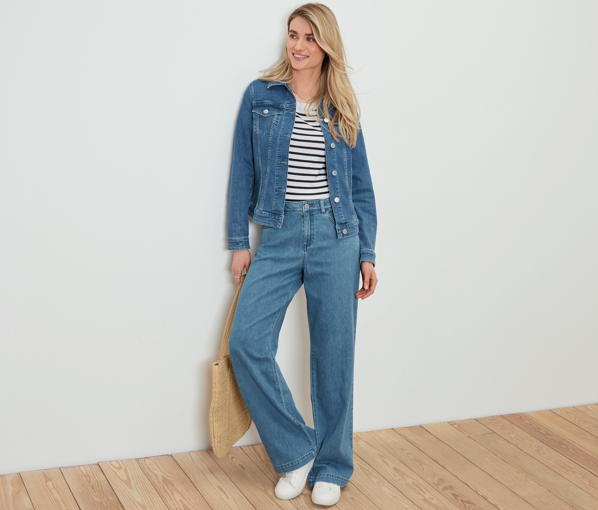 Blonde Frau lehnt an einer weißen Wand und trägt eine Jeansjacke, einen gestreiften Pullover, Palazzo-Jeans und hält einen Shopper in Bastoptik.