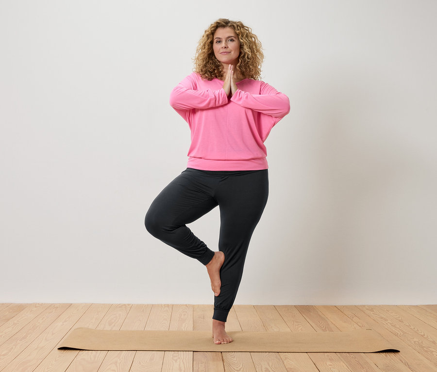 Frau mit lockigem Haar macht Yoga auf einer beigen Matte, gekleidet in ein pinkfarbenes Oberteil und graue Hosen.