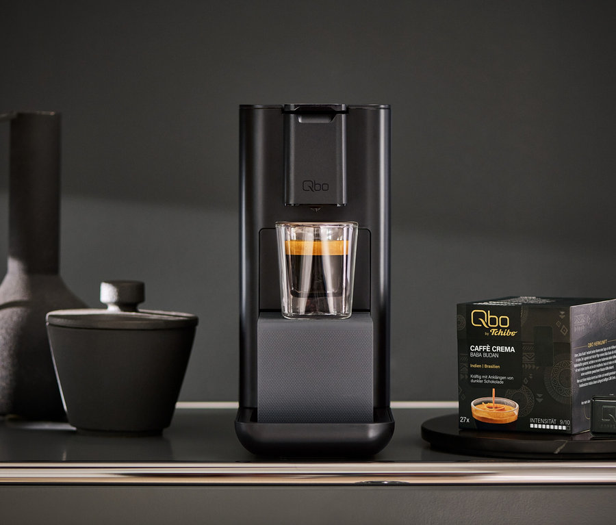 Qbo ICONIC Dark Chrome Kaffeemaschine mit Glas Tasse und Qbo Kapselpackung auf einem Tisch.