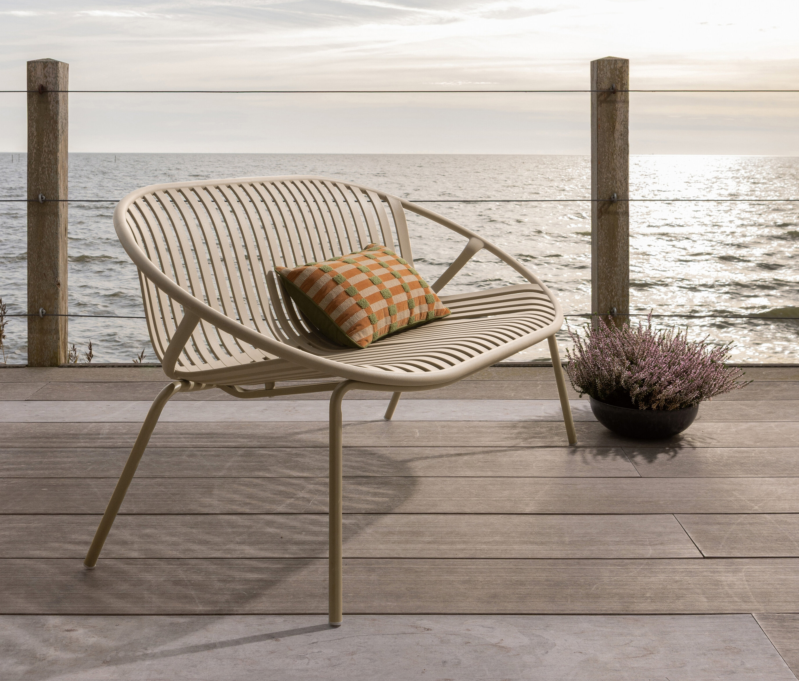 Ein beiger Outdoor-Sessel mit einem karierten Kissen und einer Topfpflanze auf einer Holzterrasse mit Blick auf das Meer.