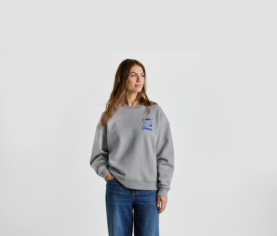 Frau mit grauem Pullover mit blauem 'Cheers'-Aufdruck und Jeans.