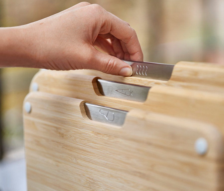 Eine Hand greift nach einem von drei Schneidebrettern aus Bambus mit silbernen Griffen, die jeweils mit Symbolen versehen sind.