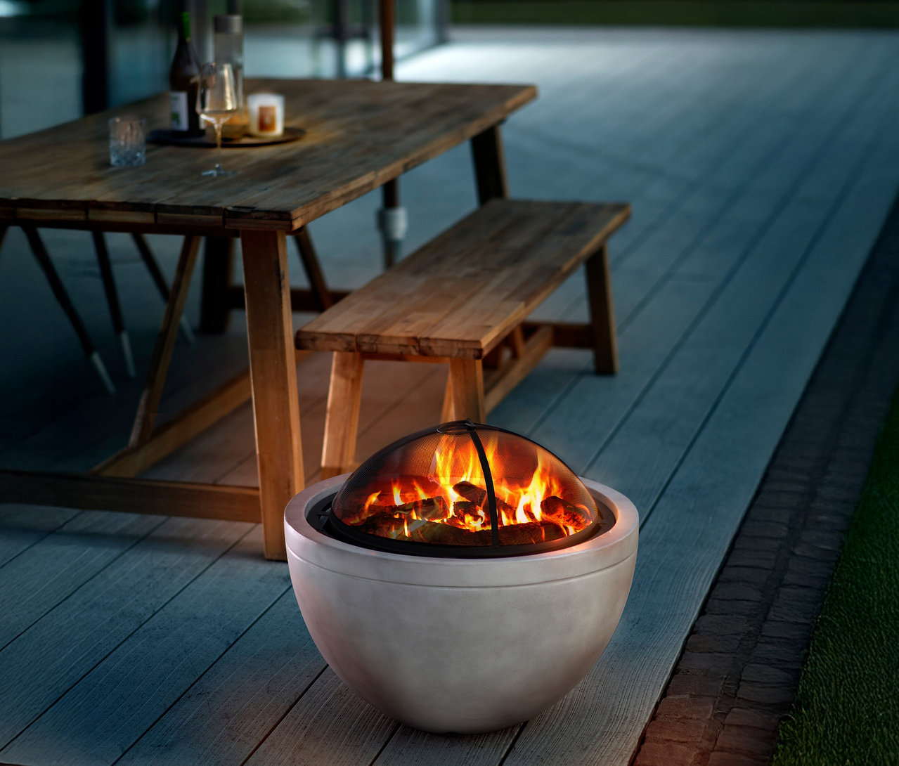 Feuer brennt in einer grauen Feuerstelle auf einer Holzterrasse; im Hintergrund ein Holztisch mit Bank.