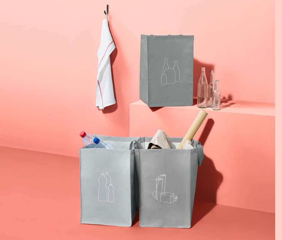 Drei graue 3 Recycling-Taschen, zwei davon gefüllt, stehen auf dem Boden, eine auf einem Podest.