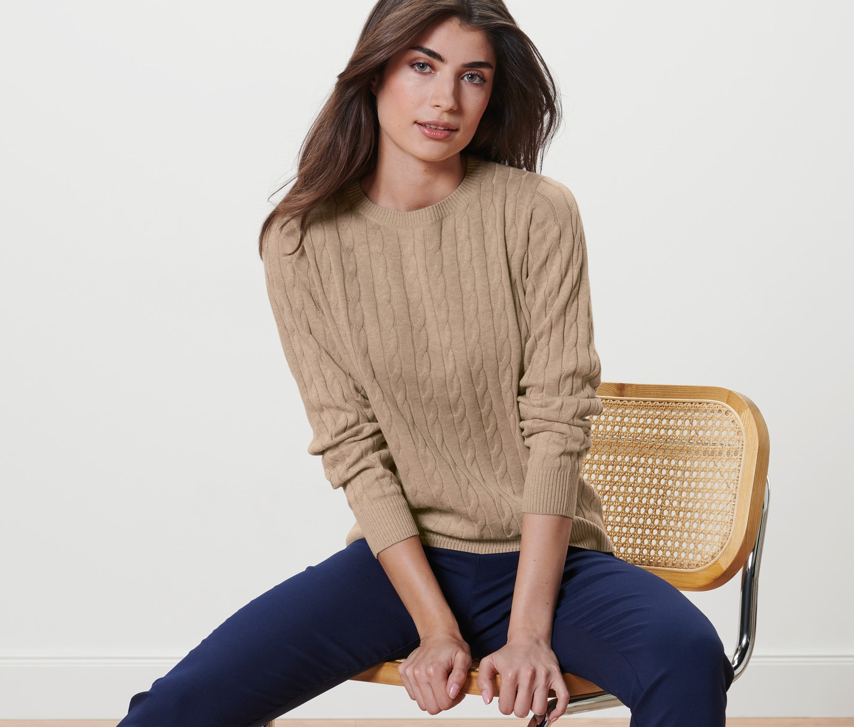 Frau sitzt auf einem Stuhl und trägt einen creme melierten Zopfstrickpullover.