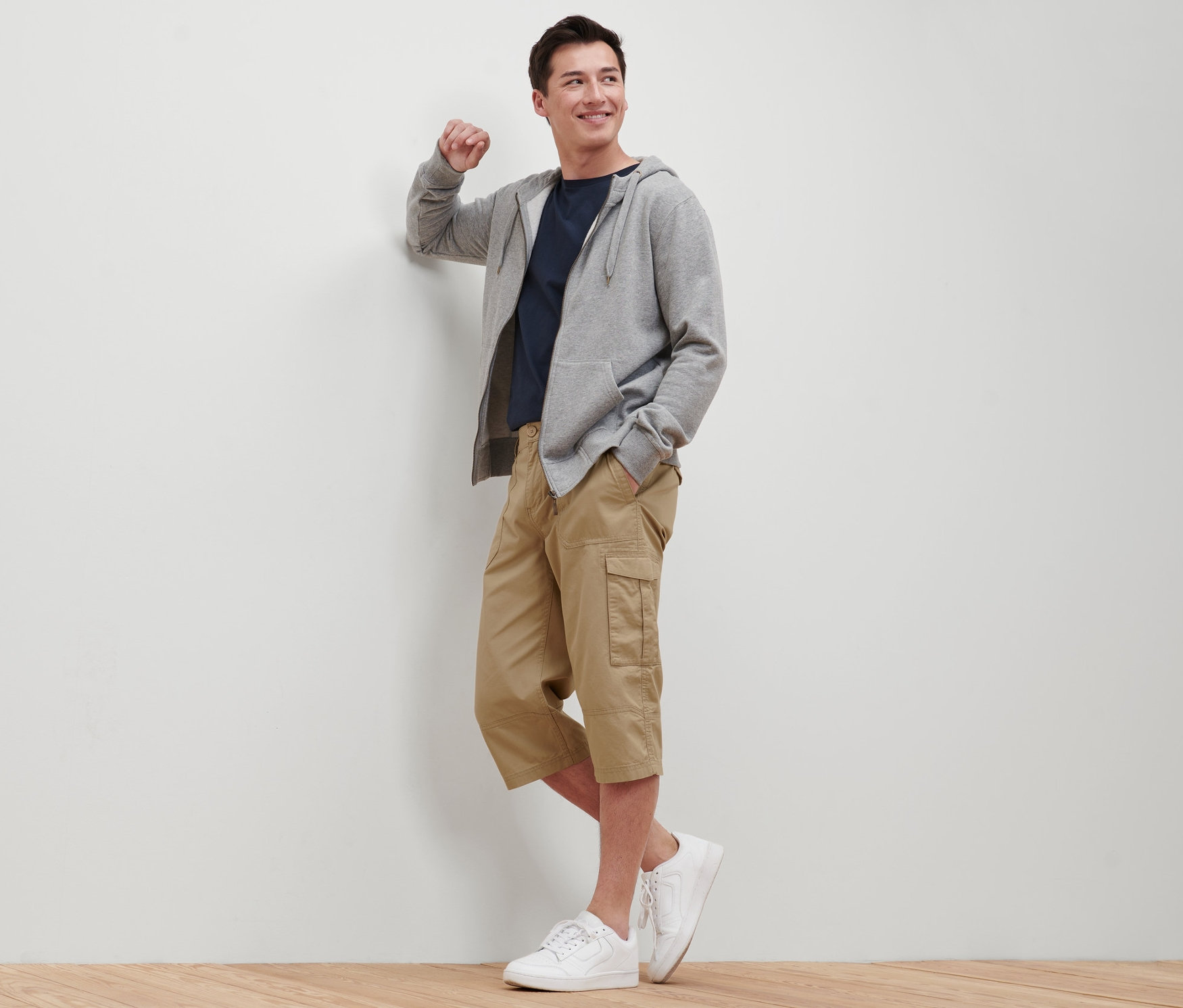 Mann lehnt an Wand, trägt beige Cargo-Bermuda in 3/4-Länge, T-Shirt, Kapuzenpullover und Turnschuhe.