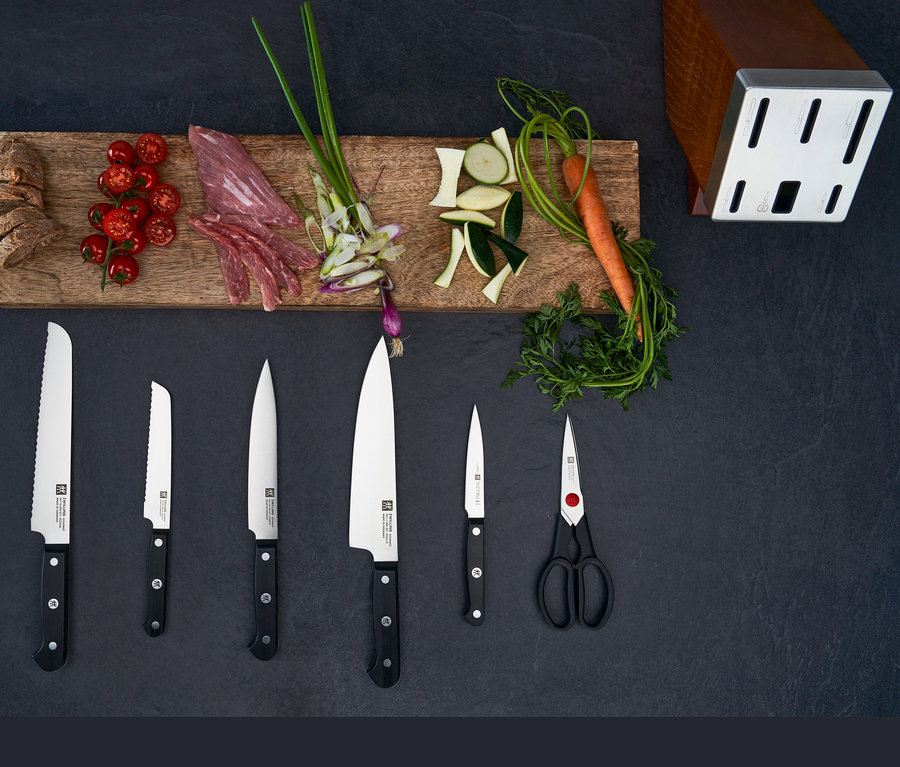 Auf dunklem Grund liegen ein Holzbrett mit geschnittenem Brot, Tomaten, Fleisch, grüne Zwiebeln, geschnittene Zucchini und Karotten. Darunter liegen fünf Messer und eine Schere. Rechts steht ein ZWILLING Selbstschärfender Messerblock »Gourmet«.