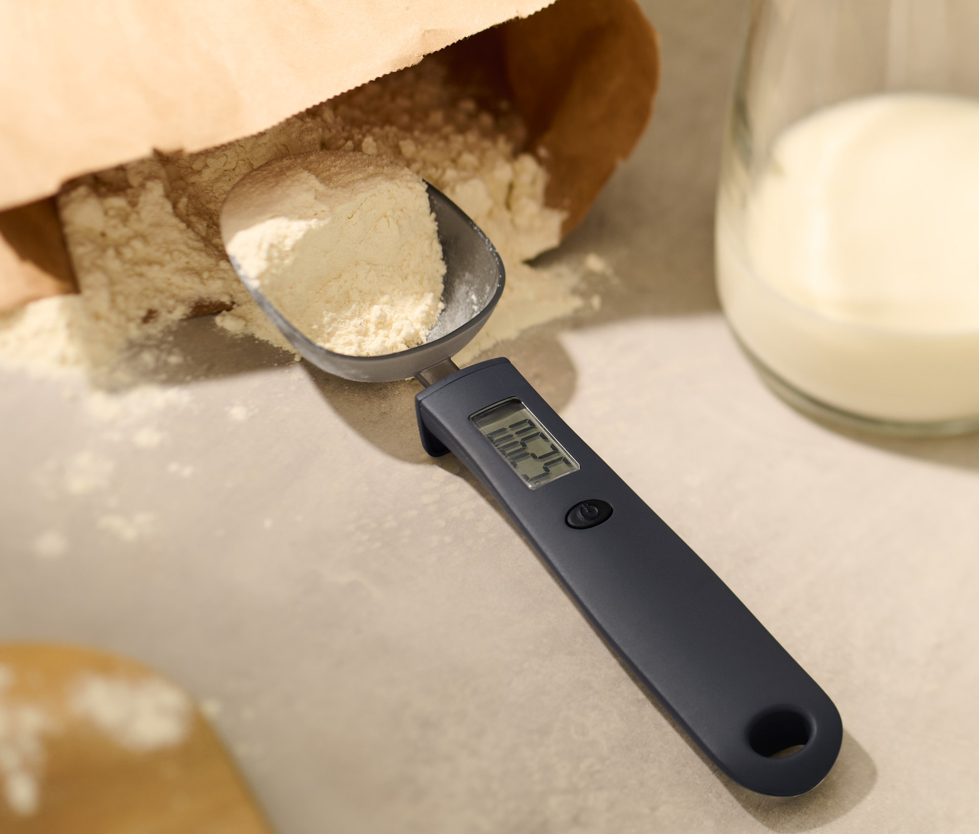 Mehl in digitaler Löffelwaage neben einem Glas Milch.