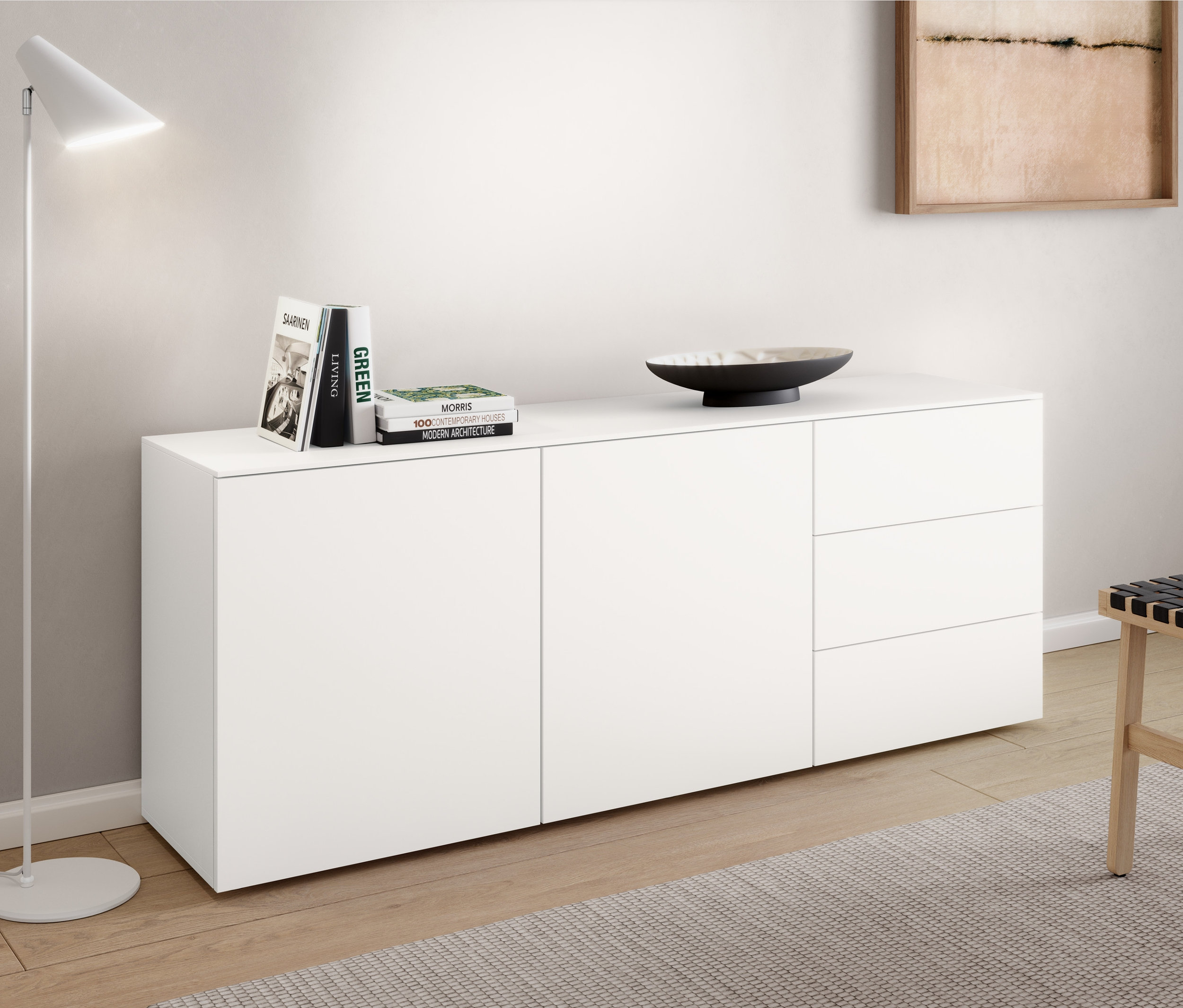 Eine weiße Metall-Stehleuchte »Cale« steht neben einem Sideboard. Auf dem Sideboard liegen Bücher und eine Schale. Neben dem Sideboard steht ein Hocker mit Geflecht.