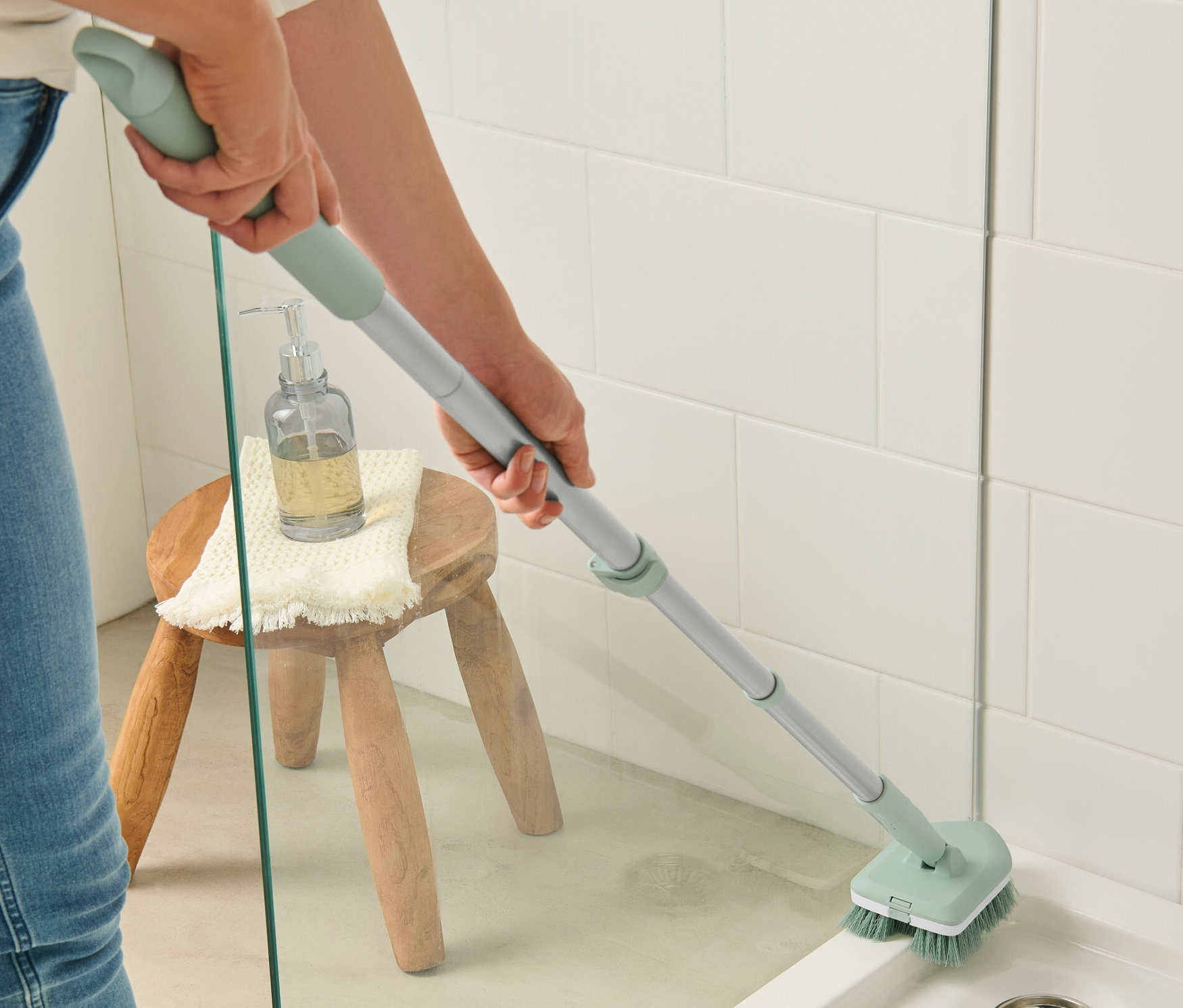 Eine Frau putzt eine Dusche mit einem Fliesen-und-Badschrubber. Auf einem Hocker stehen ein Seifenspender und ein Handtuch.