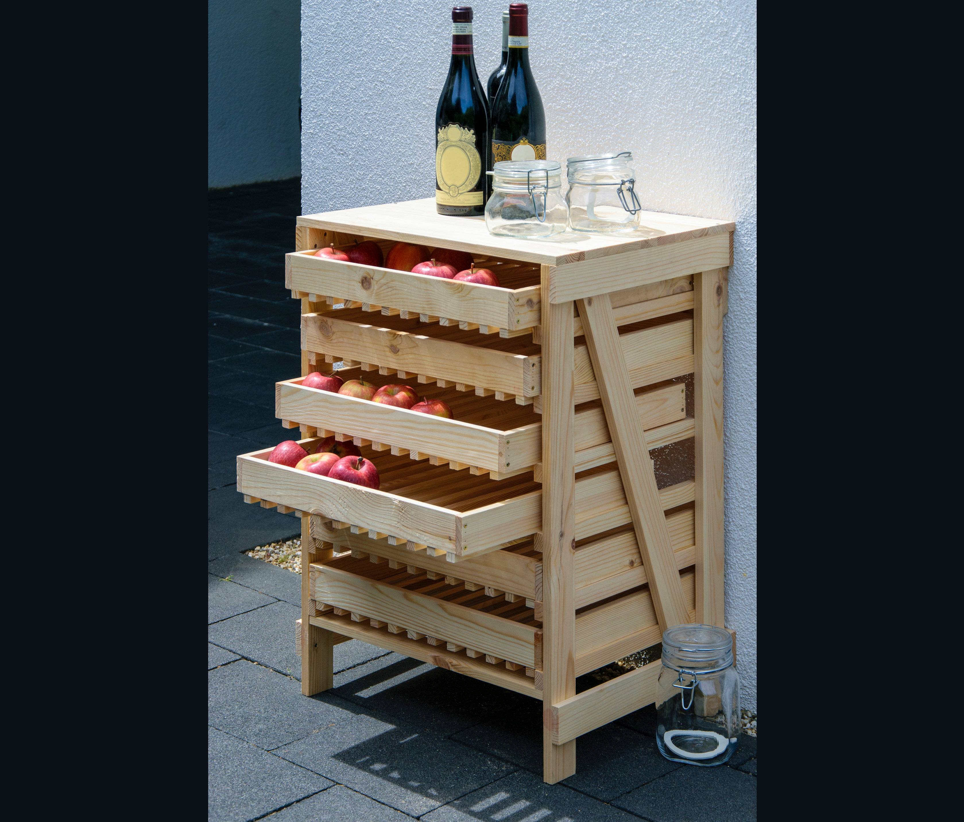 Ein Dobar Vorratsregal aus Holz mit drei Schubladen voller roter Äpfel, auf der Oberseite zwei Weinflaschen und zwei Gläser.