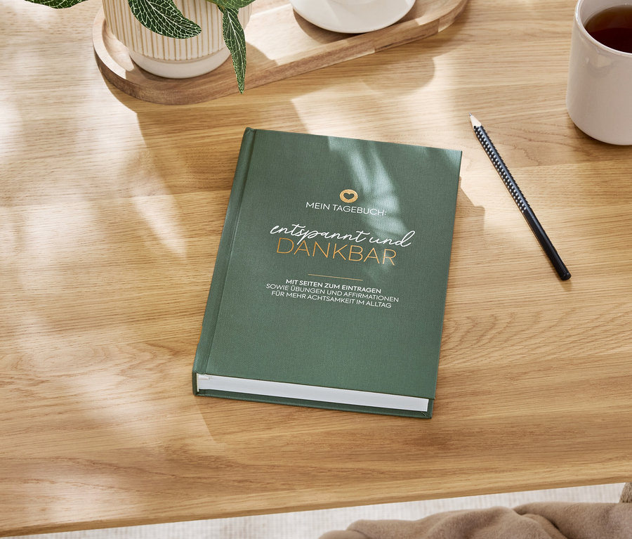 Ein grünes Notizbuch 'Entspannt und dankbar' liegt auf einem Holztisch neben einem Stift und einer Tasse mit Getränk.