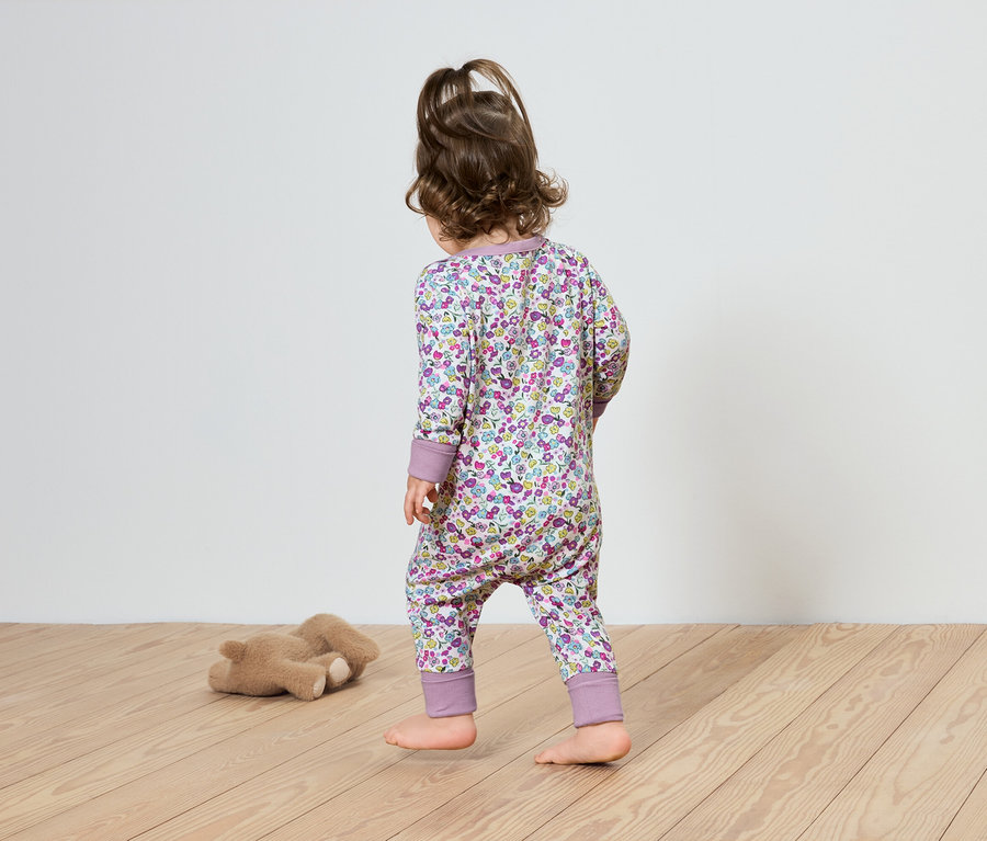 Ein Kind in einem geblümten Overall geht auf einem Holzboden in Richtung eines Teddybären.