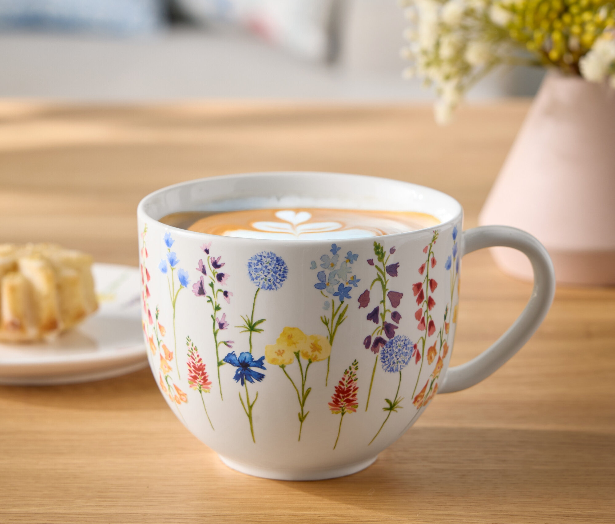 Blumenbecher mit Kaffee und Latte-Art-Herz auf einem Holztisch neben einem Teller mit Gebäck und einer Blumenvase.