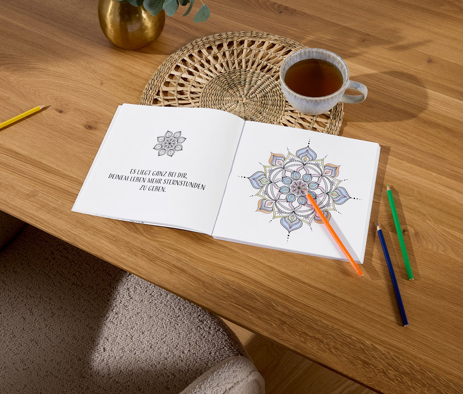 Ein geöffnetes Malbuch mit einem Mandala liegt auf einem Holztisch neben einer Tasse Tee und bunten Stiften.