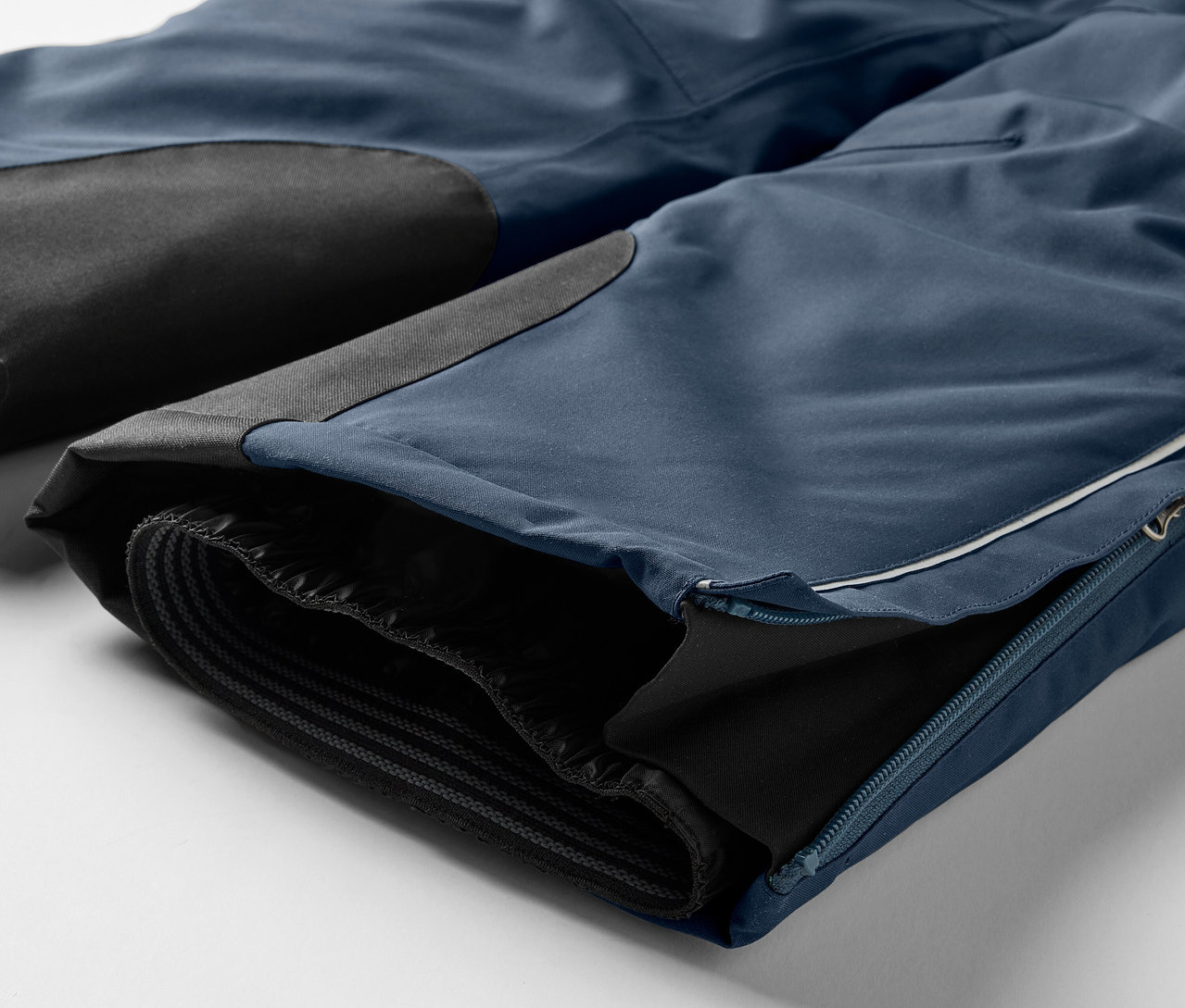 Detail einer blauen Hose mit schwarzen Knieeinsätzen und einem Reißverschluss am Bein.