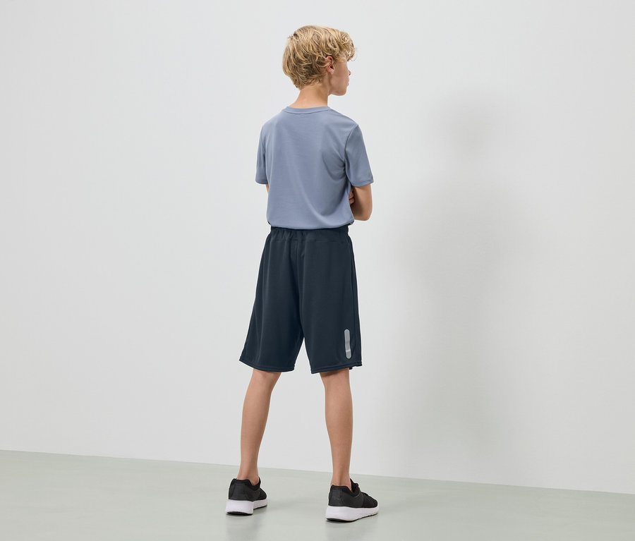 Ein Jugendlicher mit blonden, lockigen Haaren steht mit dem Rücken zur Kamera, trägt ein blaues T-Shirt, marineblaue Shorts und schwarze Turnschuhe.