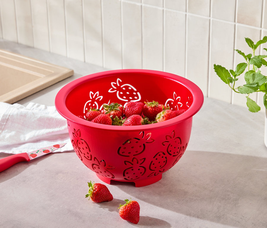 Eine rote Schüssel mit Erdbeermuster ist mit Erdbeeren gefüllt, daneben liegt ein Messer mit Erdbeermuster.