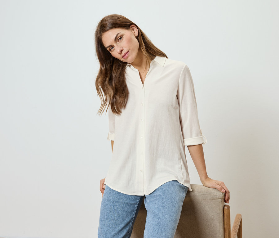 Frau mit gewelltem Haar, trägt ein helles Hemd und Jeans, lehnt an einem Stuhl.