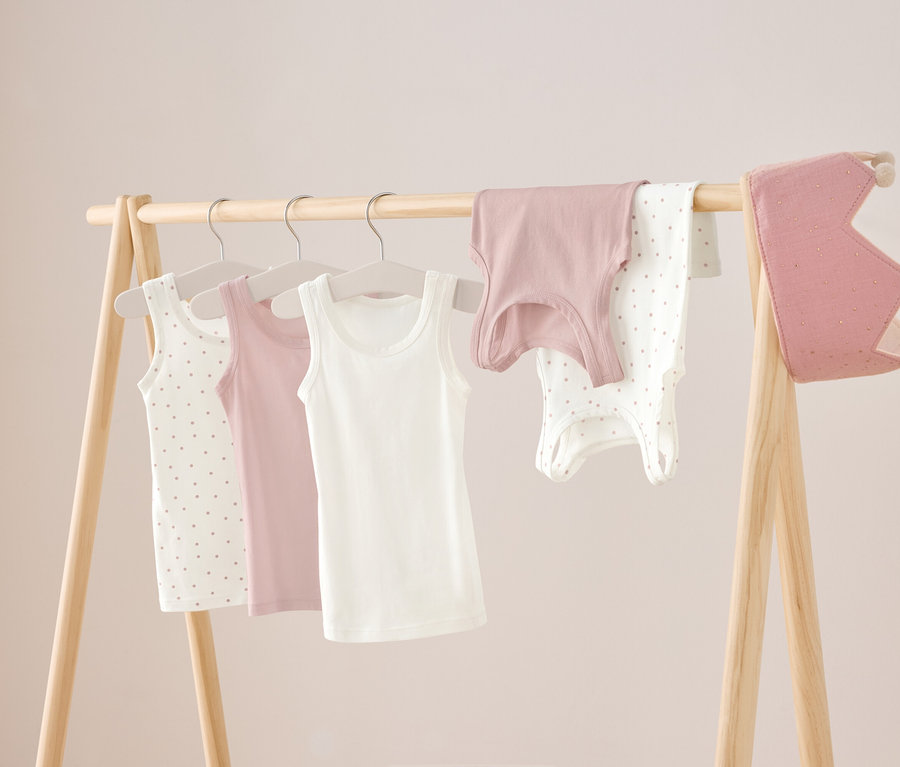 Auf einem Holzständer hängen drei Unterhemden in Weiß, Rosa und Weiß mit Punkten, sowie ein rosa Hemd und ein weißer Body mit Punkten. Rechts hängt eine rosa Krone.
