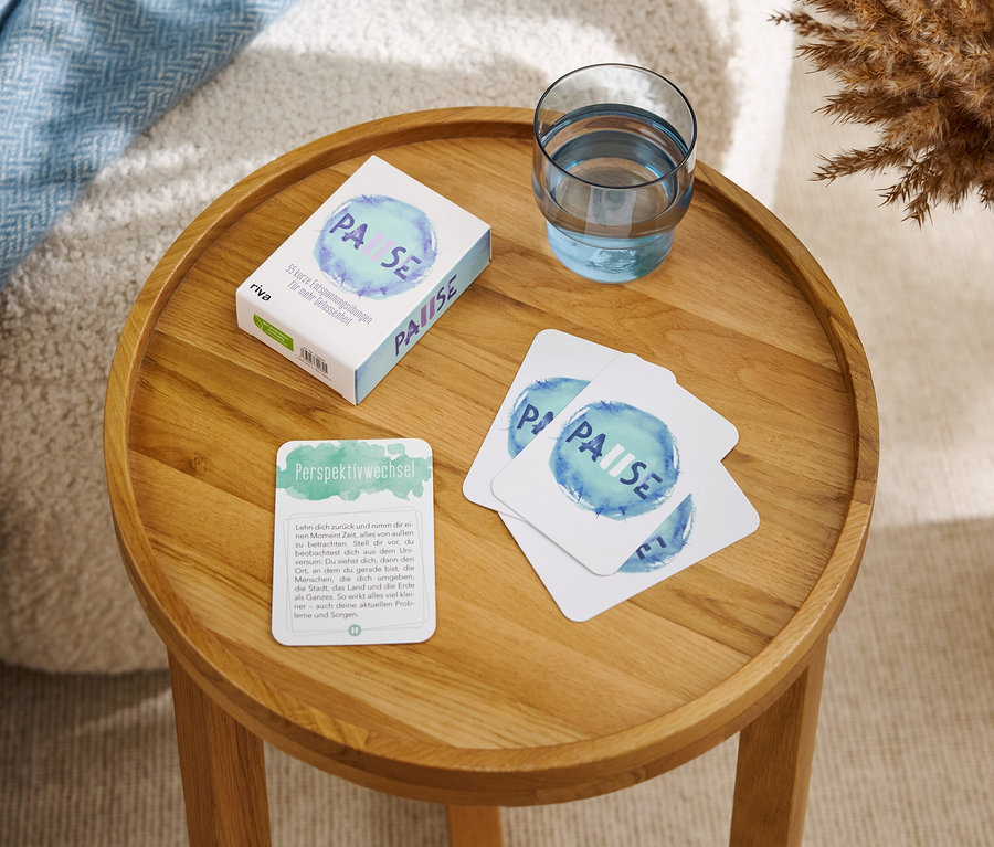 Auf einem runden Holztisch liegen eine Schachtel und Karten mit der Aufschrift "PAUSE" sowie ein Glas Wasser.