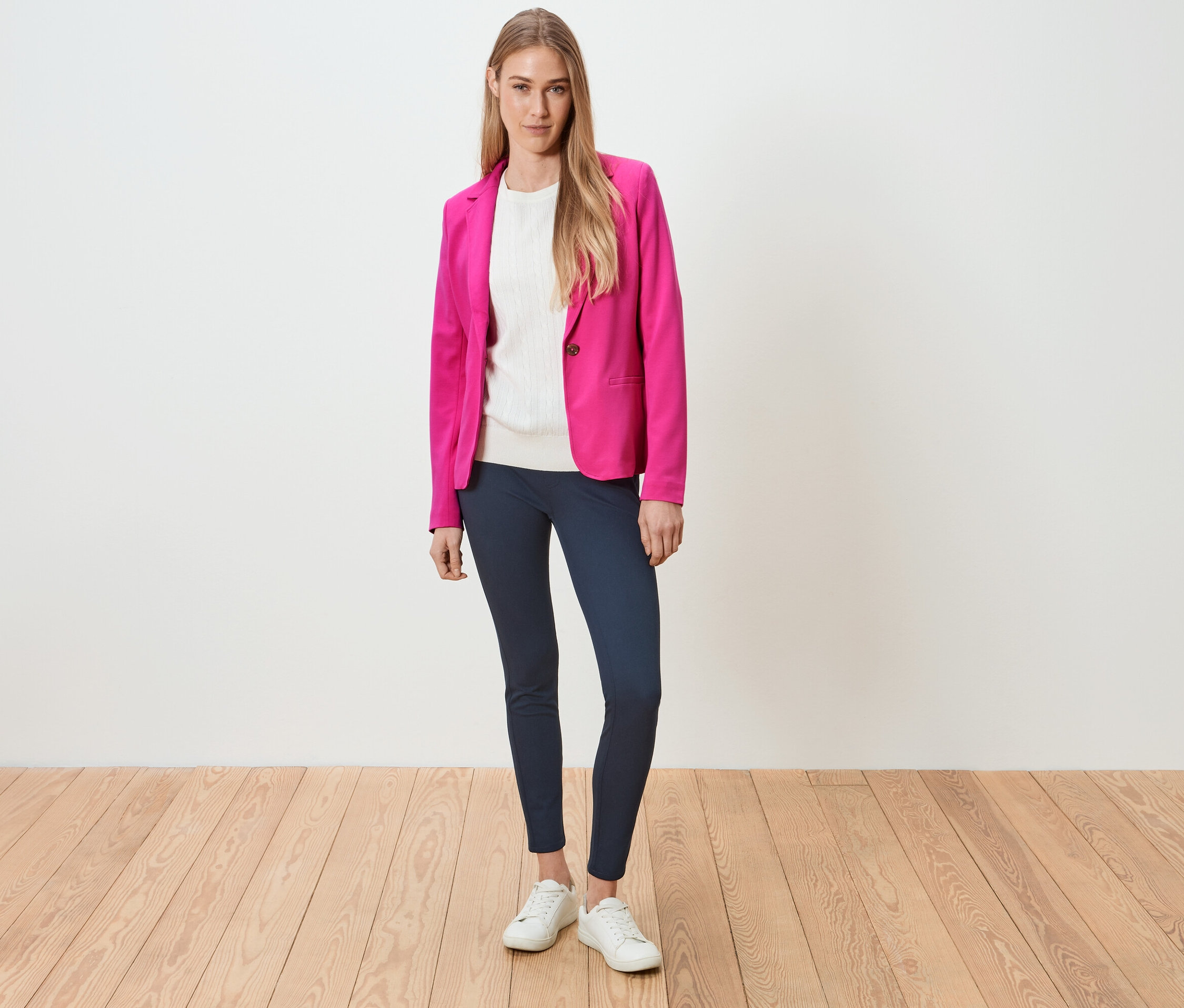 Frau steht und trägt einen pinken Blazer, ein weißes Oberteil, blaue Leggings und weiße Turnschuhe.