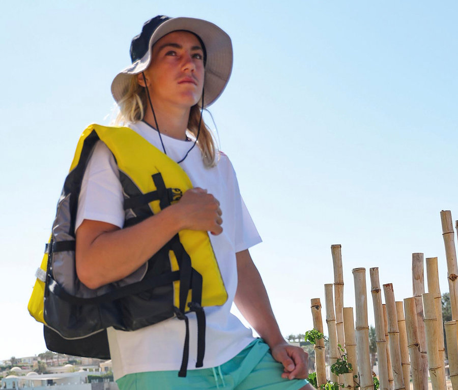 Ein Mann mit Hut und Schwimmweste steht vor Bambusstangen. Er trägt ein weißes T-Shirt und türkisfarbene Shorts.