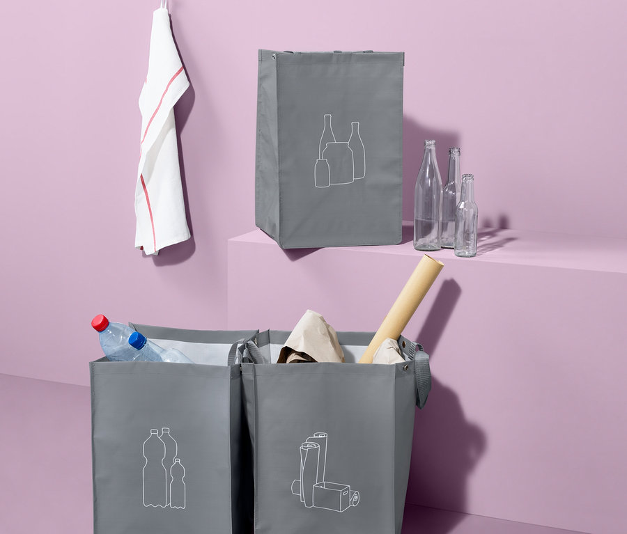 Drei graue Recycling-Abfall-Sortiertaschen mit Aufdruck stehen vor einem lila Hintergrund. Die Taschen sind mit Wertstoffen gefüllt.