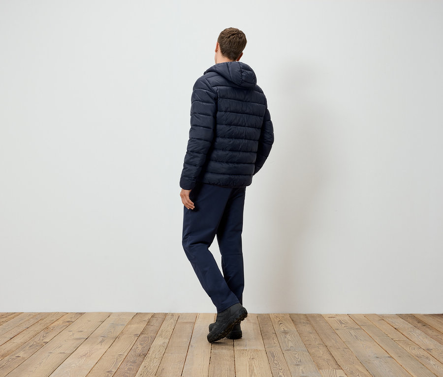 Mann von hinten steht auf einem Holzboden und trägt eine dunkelblaue Kapuzenjacke und Hose.