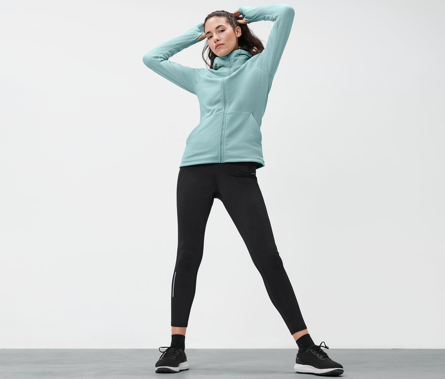 Frau posiert in hellblauer Thermojacke, schwarzen Leggings und Schuhen.