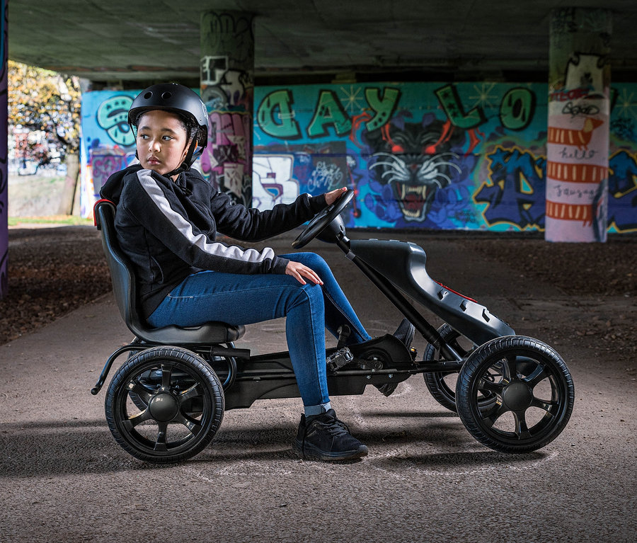 Ein Mädchen mit Helm sitzt auf einem schwarzen Xootz Gokart "Venom" unter einer Brücke mit Graffiti.