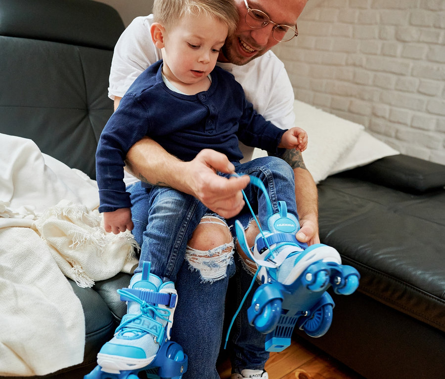Vater hält Sohn auf dem Schoß und hilft ihm beim Üben auf zyanfarbenen HUDORA My First Quad Rollschuhen.