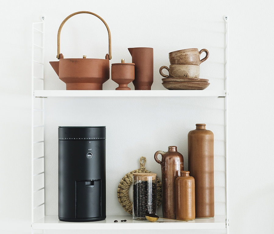 Auf einem weißen Regal stehen eine braune Keramikkanne, eine Tasse und eine Wilfa Kaffeemühle »Svart Uniform+«.