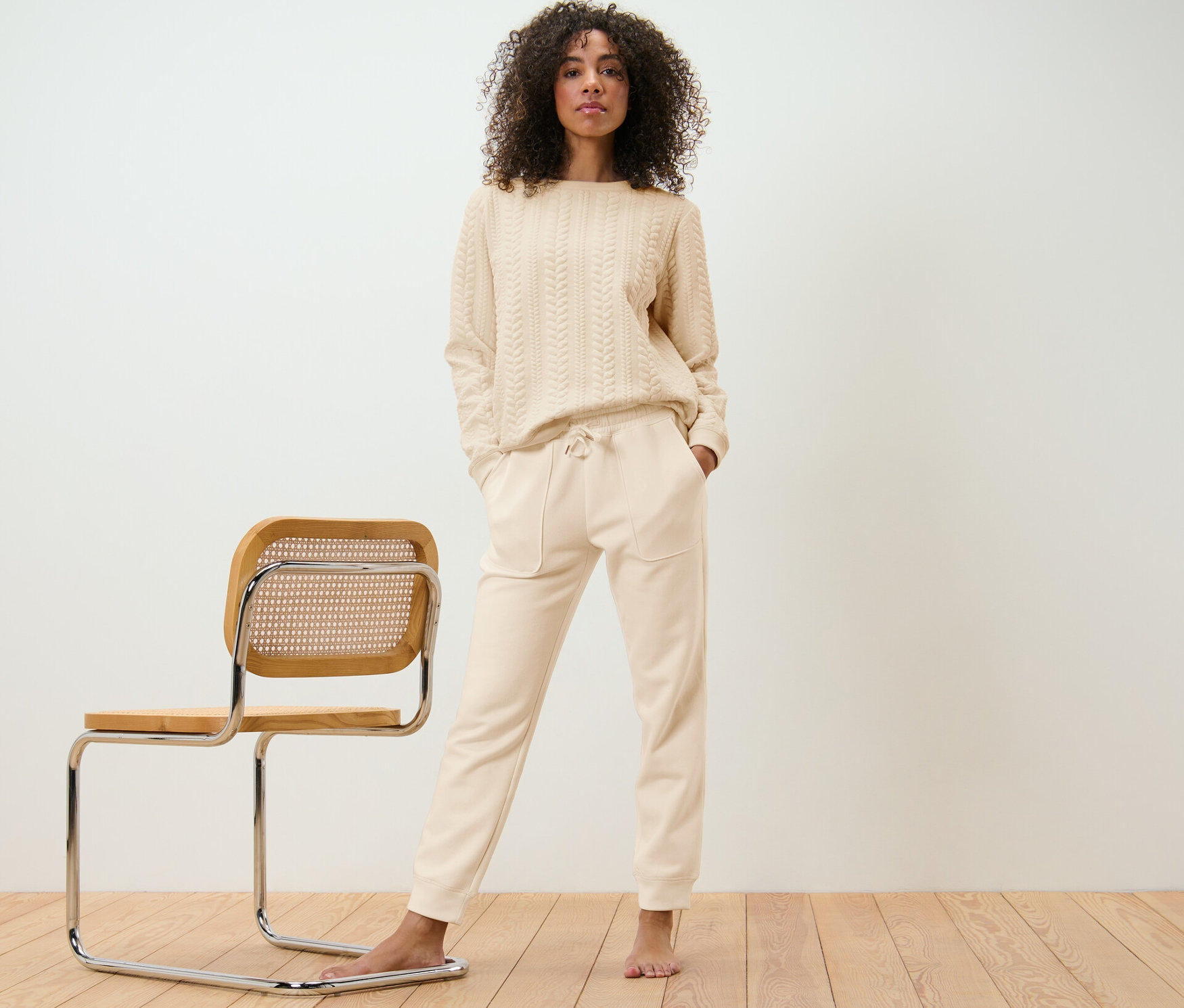 Frau posiert in beigem Hausanzug neben einem Stuhl.