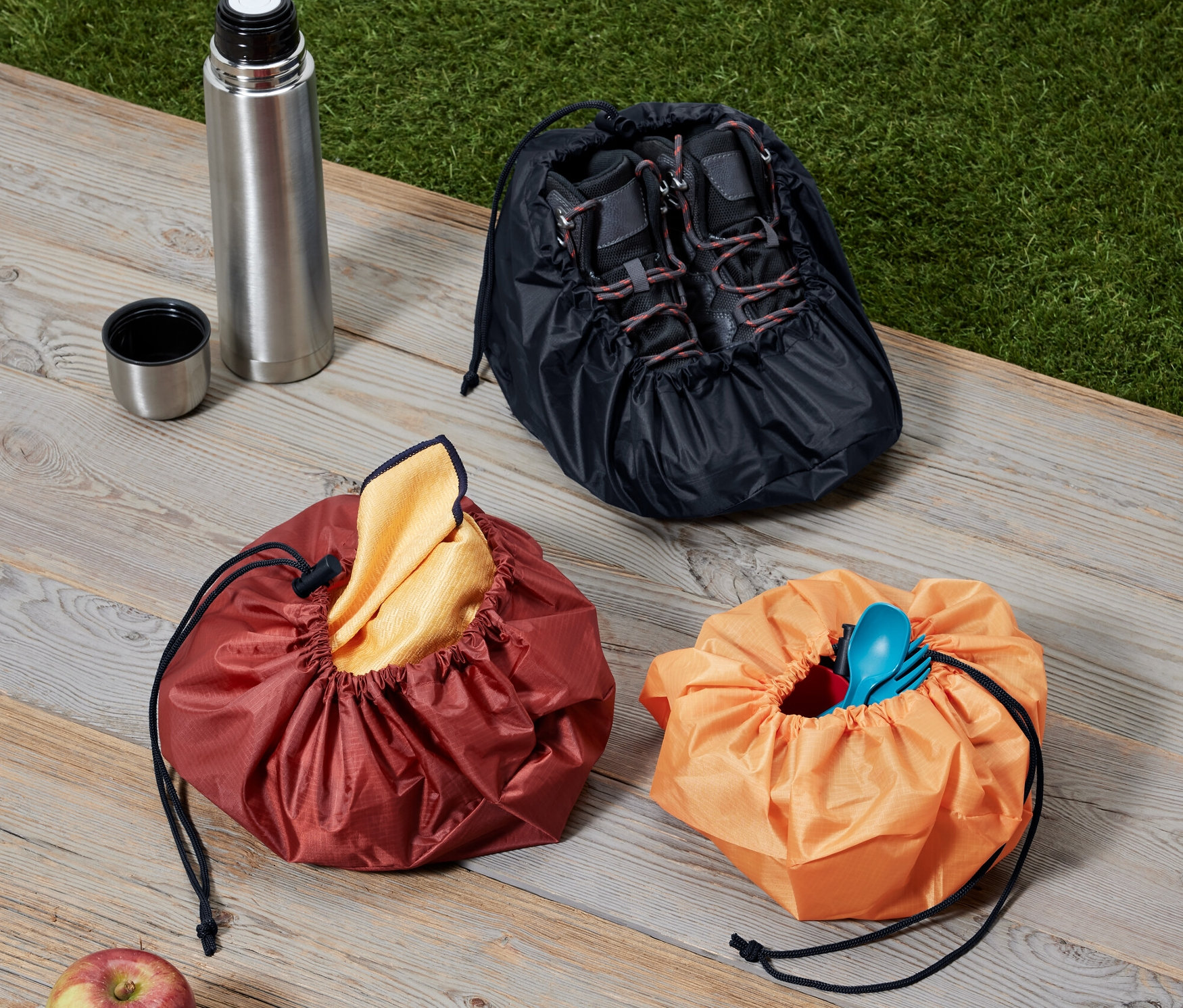 Drei Packsäcke, eine Thermoskanne und ein Apfel liegen auf einer Holzoberfläche.