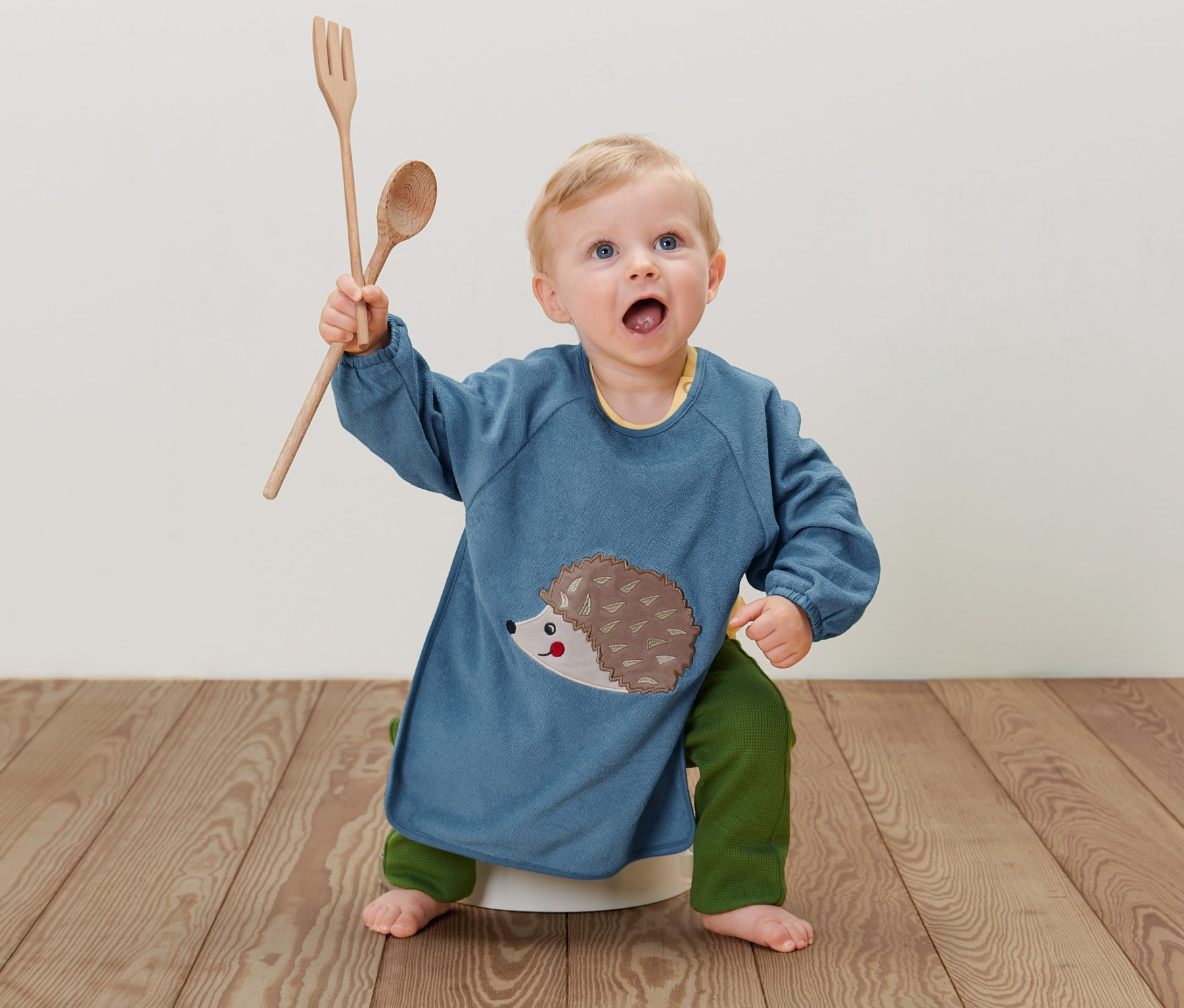 Kleinkind sitzt mit einem blauen 2 Baby-Ärmellätzchen mit Igel drauf auf dem Boden und hält einen Holzlöffel und eine Gabel hoch.