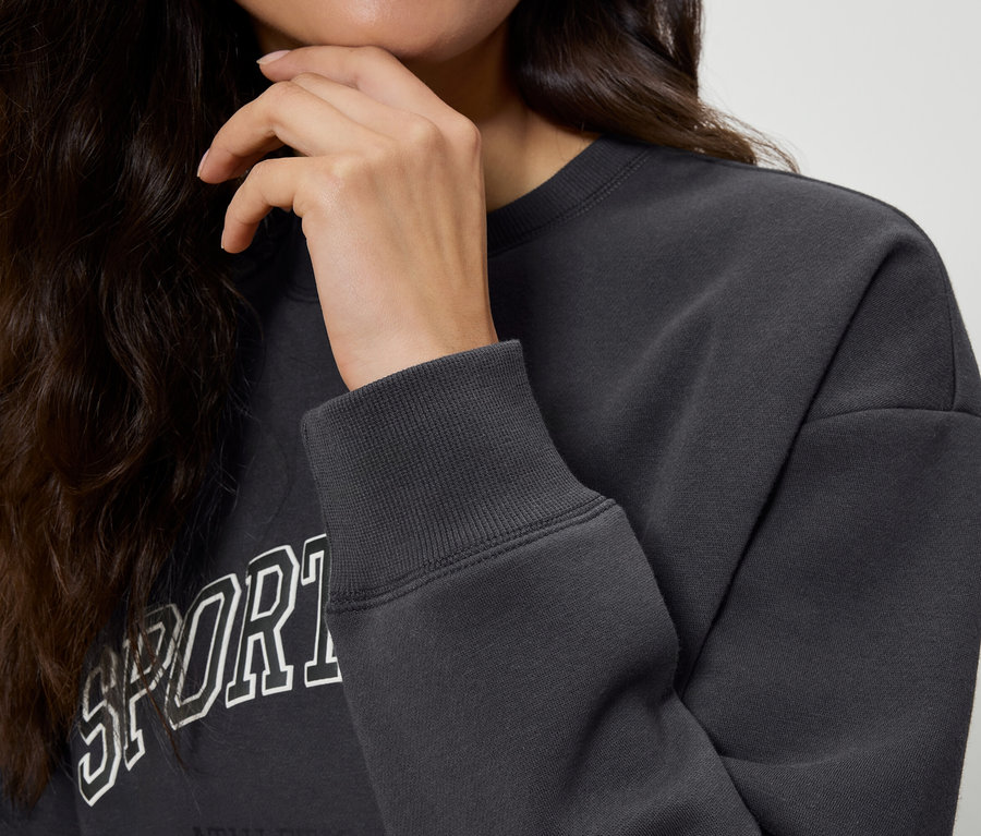 Nahaufnahme einer Frau mit gewelltem, dunklem Haar, die ihr Kinn berührt und ein dunkelgraues Sweatshirt mit der Aufschrift "Sport" trägt. Detailaufnahme des Ärmels des Sweatshirts.