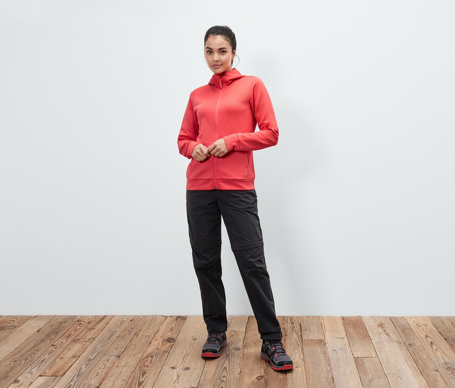 Eine Frau posiert in einer korallenroten Kapuzenjacke, schwarzen Hosen und Schuhen auf einem Holzboden.