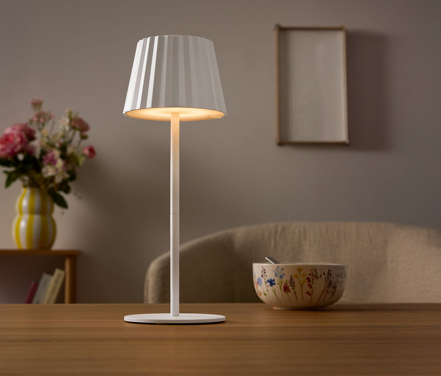 Auf einem Tisch steht eine weiße Lampe mit einem plissierten Lampenschirm. Im Hintergrund befinden sich eine Blumenvase und eine Schüssel.