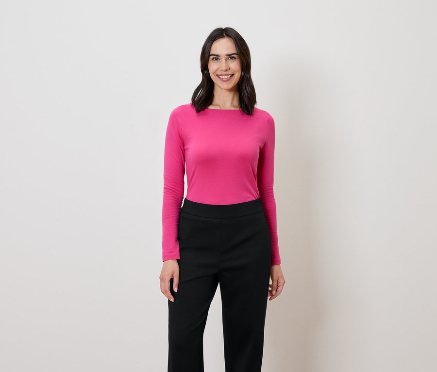 Frau mit dunklen Haaren steht und lächelt, sie trägt eine pinke Langarmbluse und eine schwarze Hose.