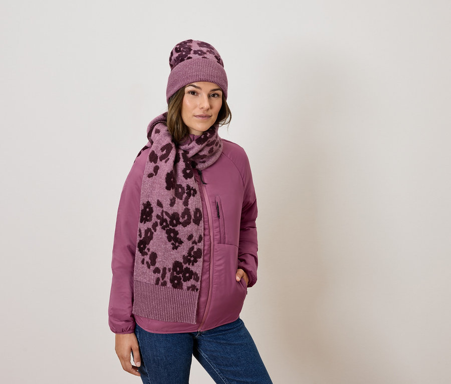 Frau trägt eine pinke Jacke, Jeans, einen Schal und eine Mütze mit Blumenmuster.